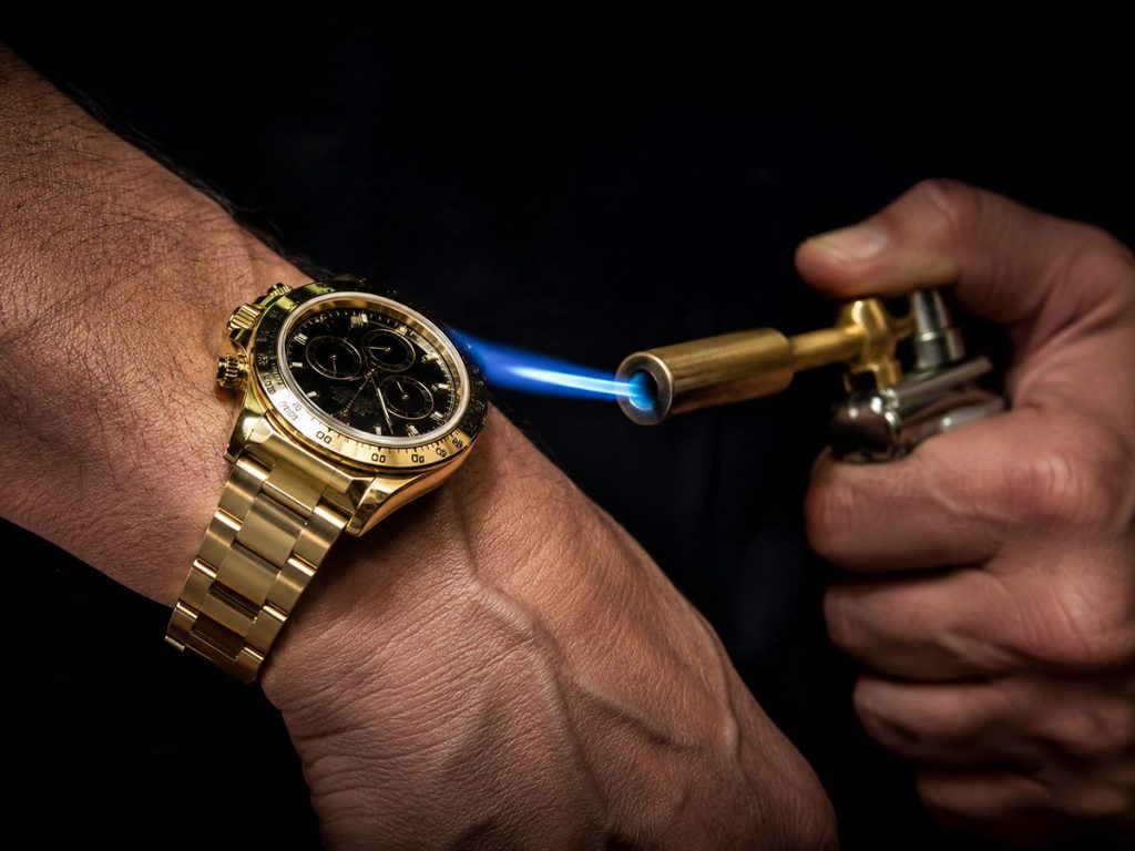 Montre de luxe en or menacée par un chalumeau de bijoutier, illustrant la fonte de montres de luxe due à la hausse du prix de l'or