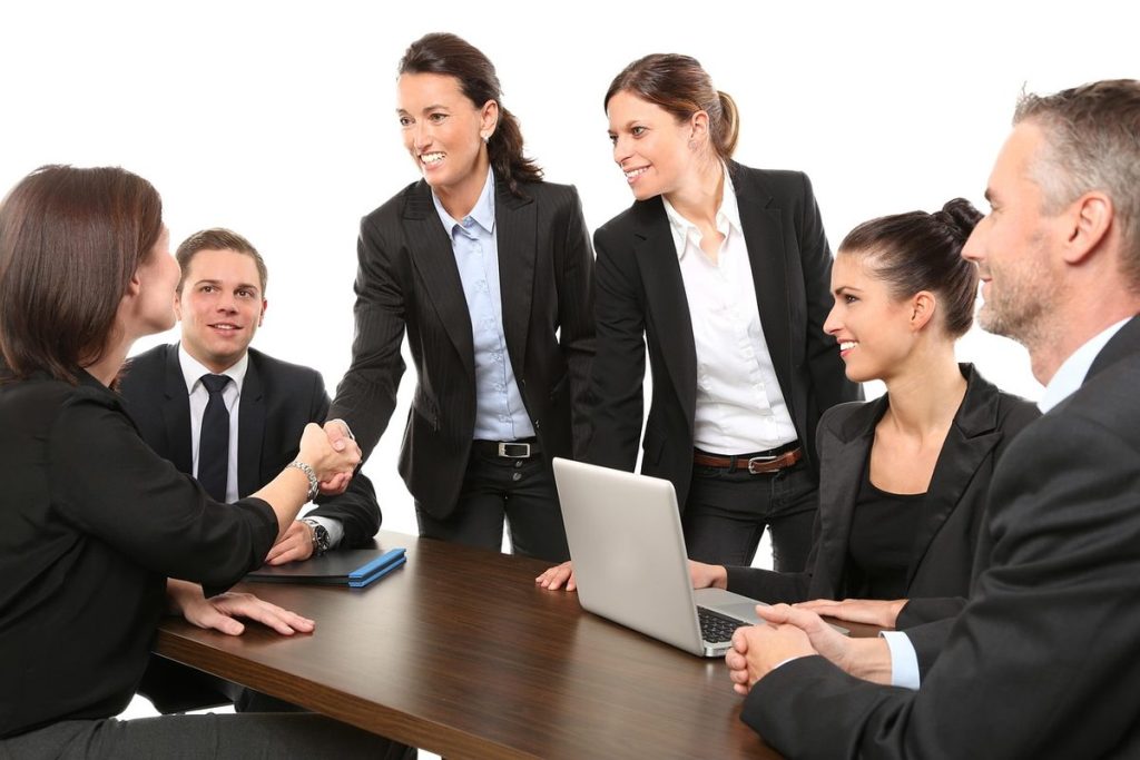 Plusieurs personnes en costume autour d'une table, deux se serrent la main, un ordinateur portable ouvert.