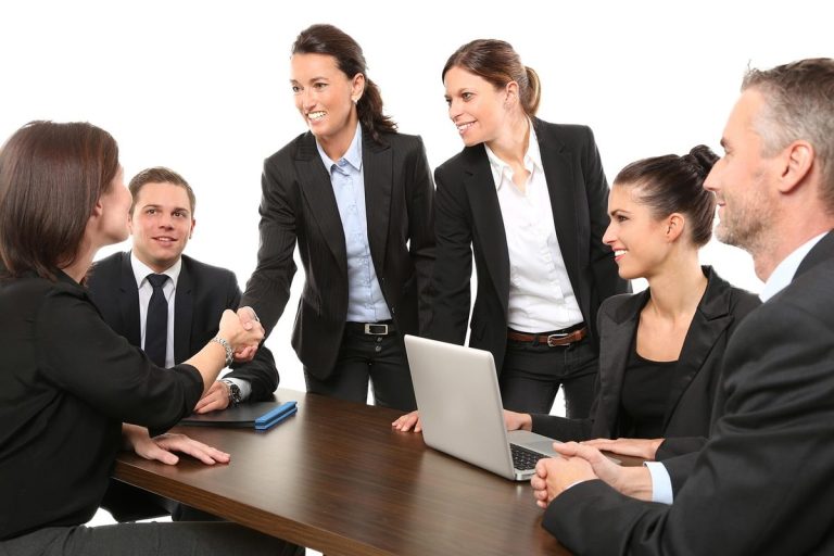 Plusieurs personnes en costume autour d'une table, deux se serrent la main, un ordinateur portable ouvert.